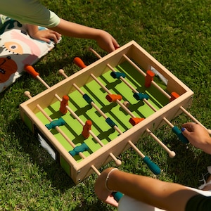 Könnte beinhalten: Ein Tischkicker aus Holz mit einem hellgrünen Spielfeld, ausgestattet mit orangefarbenen und blaugrünen Spielfiguren auf horizontalen Stangen mit passenden Griffen. Das Spiel ist rechteckig mit einem hellen Holzrahmen und einem kleinen Ball.