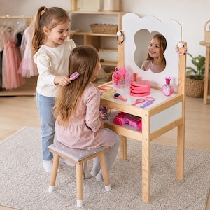 May include: A wooden vanity table with a white top, a clover-shaped mirror, and a matching stool with a gray cushion. The vanity surface holds various pink and colorful toy beauty accessories, including a hair dryer, brushes, and containers.