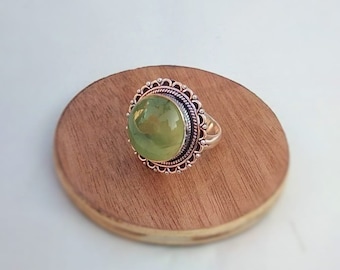May include: A silver ring with a large, round, light green gemstone. The gemstone is set in a decorative, ornate bezel with a scalloped edge. The ring is displayed on a small, round, wooden surface.