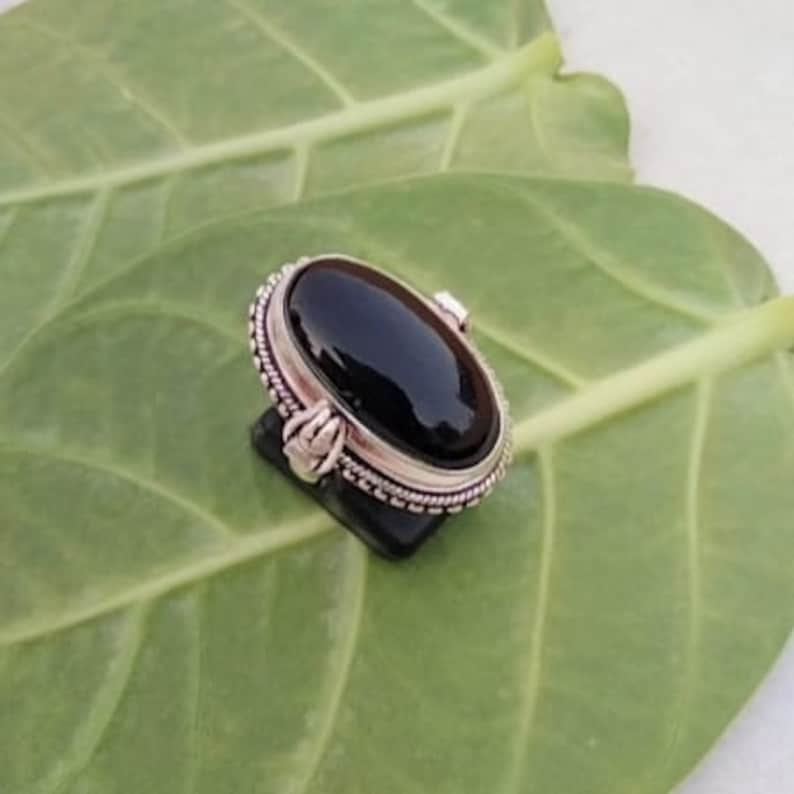 May include: A silver ring with a black oval gemstone set in a decorative bezel. The ring is on a green leaf.