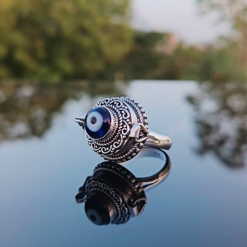 May include: A silver ring with an ornate design featuring a blue evil eye charm. The ring is on a reflective surface.