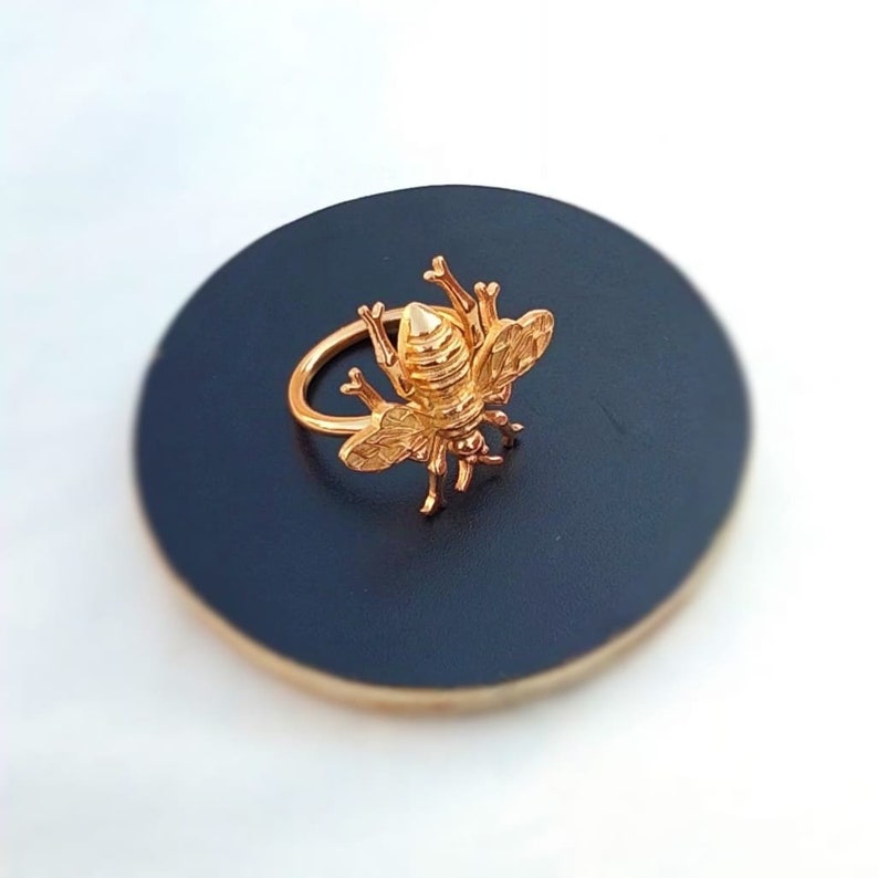 May include: A gold-colored bee ring. The ring features a detailed bee design with wings, legs, and antennae. The ring is set on a dark blue surface, creating a contrast that highlights the ring's intricate details. The ring is a piece of jewelry.