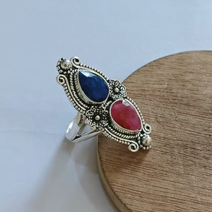 May include: A silver ring featuring two teardrop-shaped gemstones, one deep blue and the other a vibrant red. The ring has an ornate, detailed setting with floral and scrollwork designs, creating a vintage or antique aesthetic.