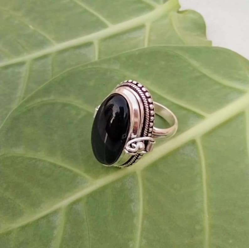 May include: A silver ring with an oval black gemstone set in a detailed silver bezel. The ring has a decorative band with small beads and a scroll design.