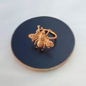 May include: A gold-colored bee ring. The ring features a detailed bee design with wings, legs, and antennae. The ring is set on a dark blue surface, creating a contrast that highlights the ring's intricate details. The ring is a piece of jewelry.