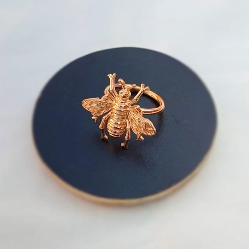 May include: A gold-colored bee ring. The ring features a detailed bee design with wings, legs, and antennae. The ring is set on a dark blue surface, creating a contrast that highlights the ring's intricate details. The ring is a piece of jewelry.