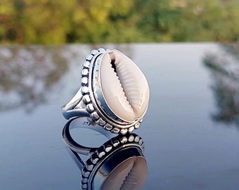 May include: A silver ring with a white cowrie shell set in a beaded bezel. The ring is reflecting on a dark surface.