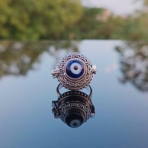 May include: A silver ring with an ornate design featuring a blue evil eye charm. The ring is reflected in a shiny surface.