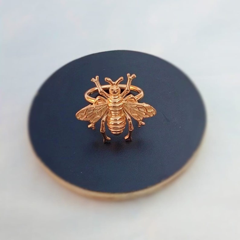 May include: A gold-colored bee ring. The ring features a detailed bee design with wings, body segments, and antennae. The ring is set on a dark blue surface, creating a contrast that highlights the ring's intricate details. The ring is a piece of jewelry.