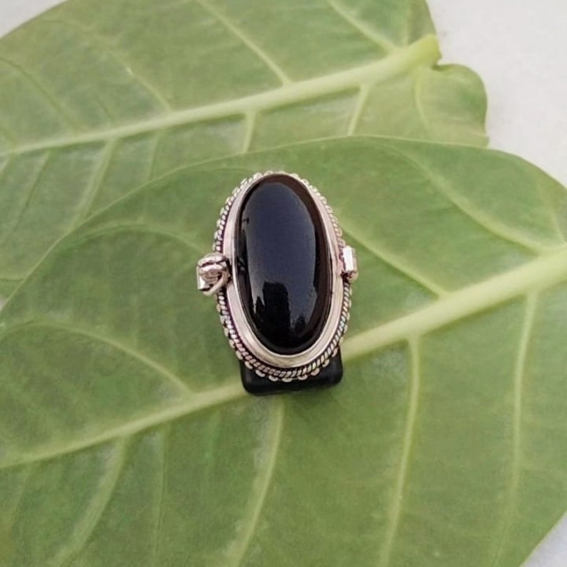 May include: A silver ring with a black oval gemstone set in a decorative silver bezel. The ring is on a green leaf background.
