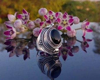 May include: A silver ring with a black oval gemstone set in a detailed filigree design. The ring is on a reflective surface with pink flowers in the background.