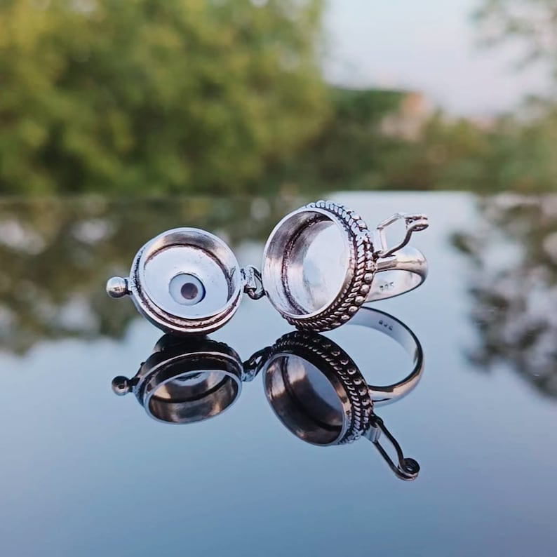 May include: A silver ring with a hinged, round compartment that opens to reveal a blue evil eye charm. The ring is decorated with a beaded design.