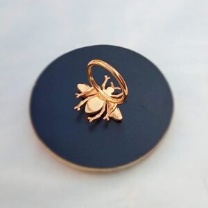 May include: Rose gold bee ring on a dark blue circular base. The ring features a detailed bee design with outstretched wings and a circular band. The bee is attached to the base, creating a unique and stylish accessory.