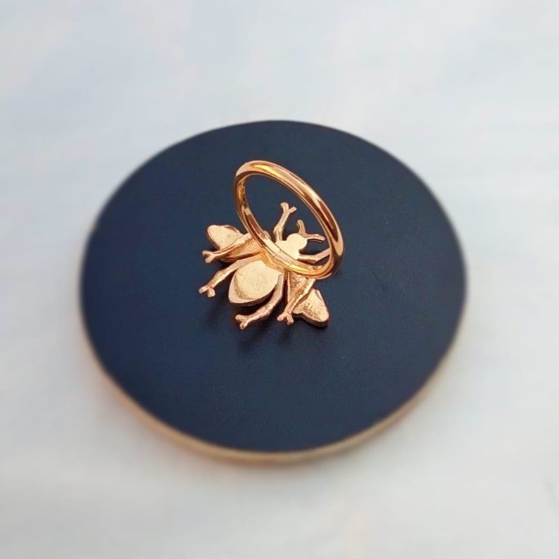May include: Rose gold bee ring on a dark blue circular base. The ring features a detailed bee design with outstretched wings and a circular band. The bee is attached to the base, creating a unique and stylish accessory.
