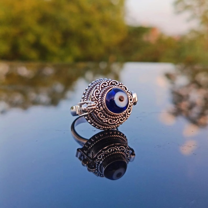 May include: A silver ring with an ornate design featuring a blue evil eye charm. The ring is on a reflective surface.
