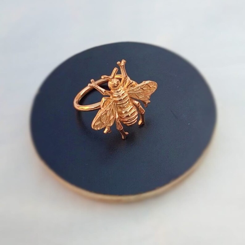May include: A gold-colored bee ring. The ring features a detailed bee design with outstretched wings and intricate body segments. The ring is set against a dark blue background, highlighting the ring's metallic sheen.