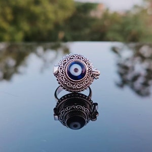May include: A silver ring with an ornate design featuring a blue evil eye charm. The ring is on a reflective surface.