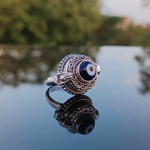 May include: A silver ring with an ornate design and a blue evil eye charm. The ring is on a reflective surface.