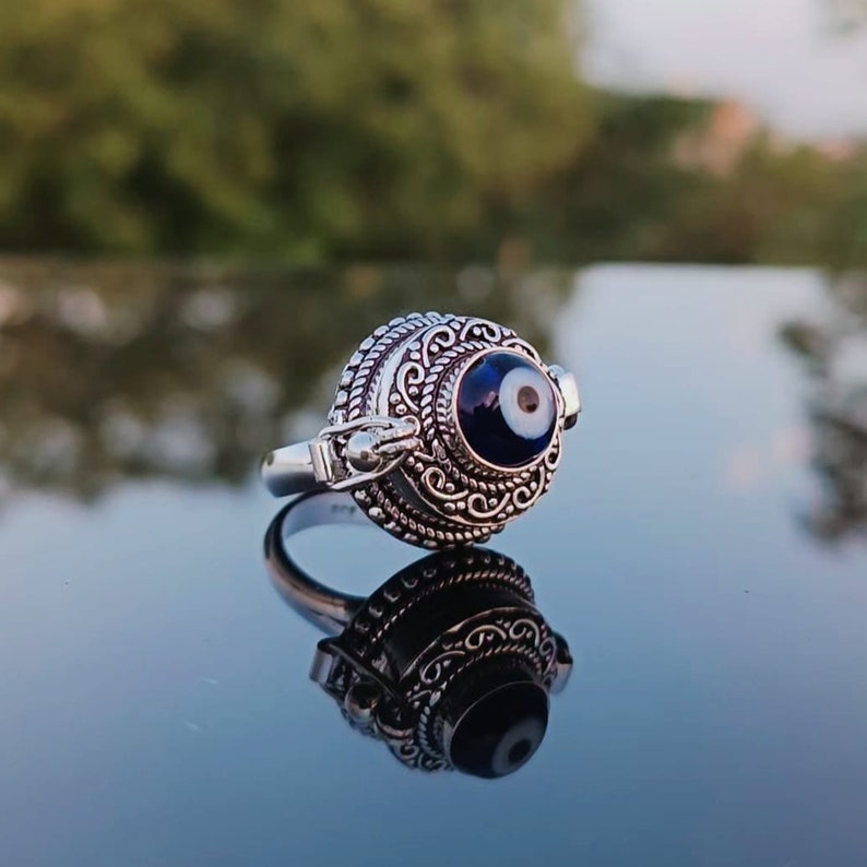 May include: A silver ring with an ornate design and a blue evil eye charm. The ring is on a reflective surface.