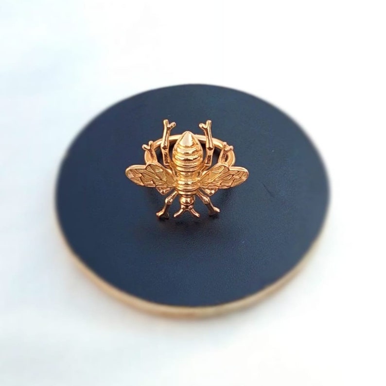 May include: A gold-colored bee-shaped decorative object. The bee is centered on a dark blue, circular surface with a gold-colored rim. The bee has detailed wings and body segments.