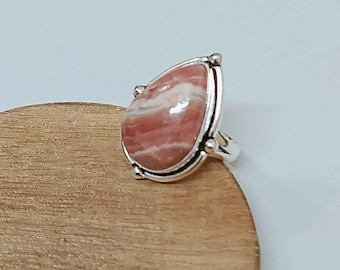 Puede incluir: Anillo de plata con una piedra preciosa grande en forma de lágrima, con bandas rosas y blancas. La piedra preciosa está engastada en un bisel de plata con detalles decorativos. El anillo se muestra sobre una superficie de madera.