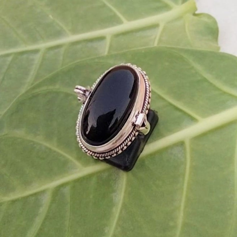 May include: A silver pendant with a black oval gemstone set in a decorative border. The pendant is on a green leaf background.