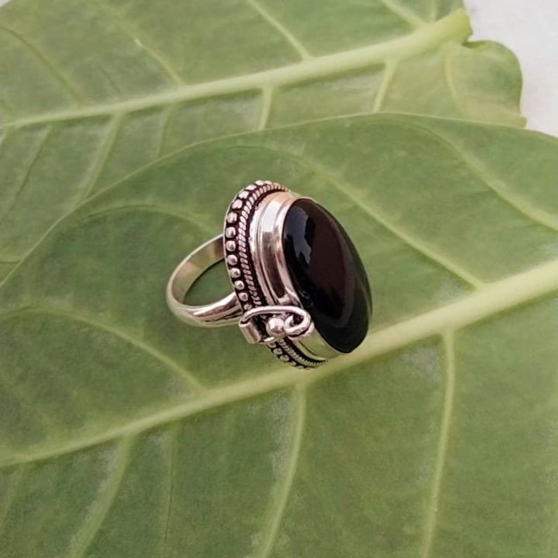 May include: A silver ring with a black oval gemstone. The ring has a decorative border and a small hinge on the side.