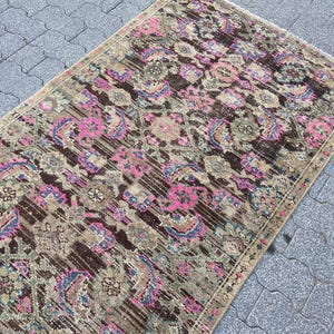 May include: Rectangular vintage rug with a distressed design. The rug features a brown, pink, and beige color palette with floral and geometric patterns. The rug is placed on a gray stone surface.