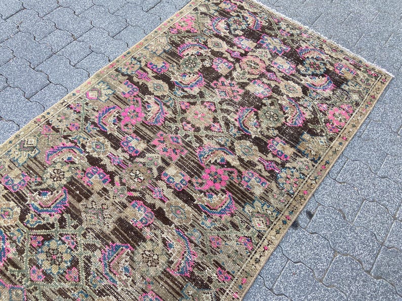 May include: Rectangular vintage rug with a distressed design. The rug features a brown, pink, and beige color palette with floral and geometric patterns. The rug is placed on a gray stone surface.
