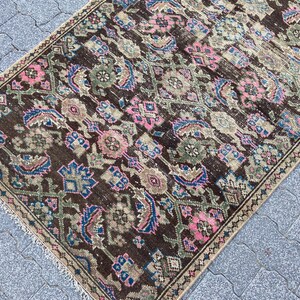 May include: A rectangular, vintage rug with an intricate, repeating pattern. The rug features a dark brown background with floral and geometric designs in shades of pink, blue, and green. The edges are finished with a fringe.
