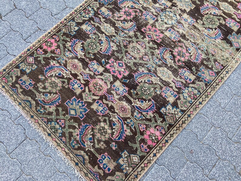 May include: A rectangular, vintage rug with an intricate, repeating pattern. The rug features a dark brown background with floral and geometric designs in shades of pink, blue, and green. The edges are finished with a fringe.