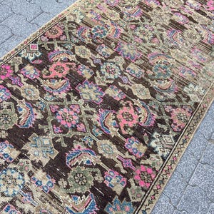 May include: A rectangular, vintage-style rug with a dark brown background and intricate floral patterns in pink, blue, and green. The rug has a distressed look, with faded areas and a beige border, suggesting it is an antique or a well-loved piece.