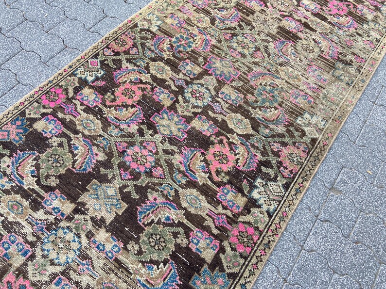 May include: A rectangular, vintage-style rug with a dark brown background and intricate floral patterns in pink, blue, and green. The rug has a distressed look, with faded areas and a beige border, suggesting it is an antique or a well-loved piece.