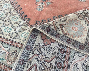May include: An antique rug with a distressed, faded appearance. The rug features a central field in a muted coral color, with intricate floral and geometric patterns in shades of beige, teal, and brown. The edges are detailed with a border.