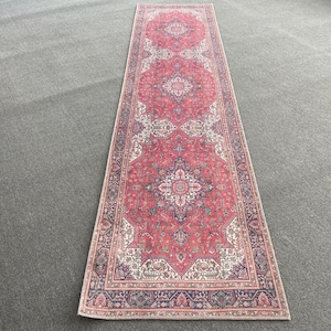 May include: A long, rectangular rug with a traditional design. The rug features a red field with intricate patterns in white, blue, and beige. The border is a darker blue with floral motifs. The rug is placed on a gray surface.