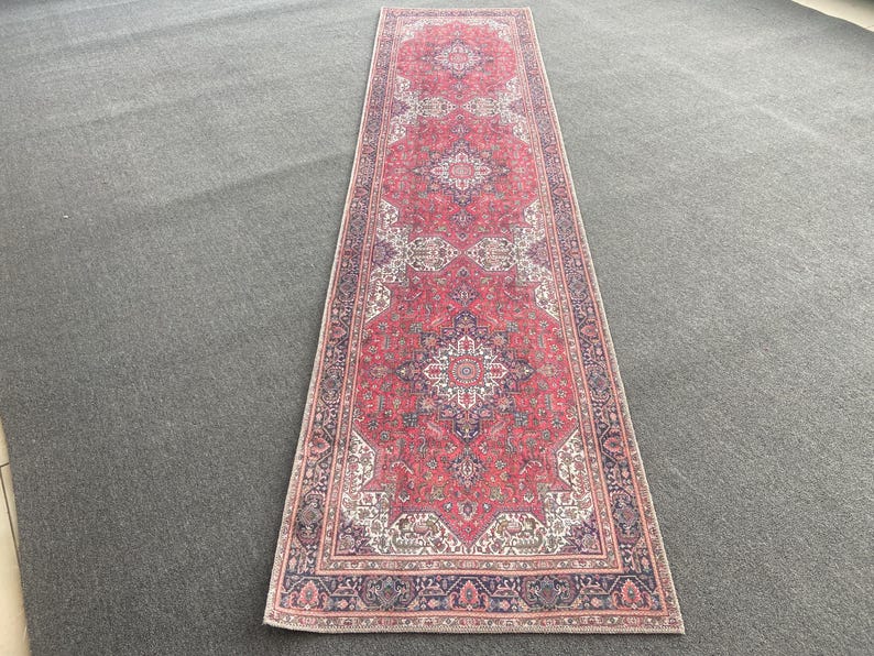May include: A long, rectangular rug with a traditional design. The rug features a red field with intricate patterns in white, blue, and beige. The border is a darker blue with floral motifs. The rug is placed on a gray surface.