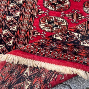 May include: A vibrant red rug with intricate geometric patterns in black, brown, and cream. The rug features a central design with repeating medallions and a detailed border. The edges are finished with a fringe.