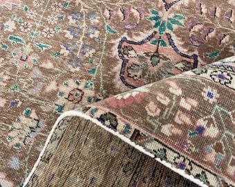 May include: A woven rug featuring a brown base with intricate floral and geometric patterns in shades of pink, teal, blue, and cream. The rug is folded over to reveal a textured, grid-like underside with a white finished edge.