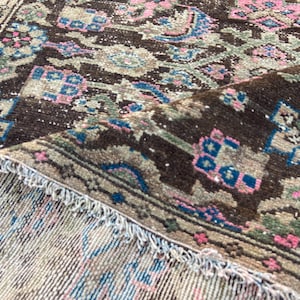 May include: Close-up of a vintage rug with a detailed, floral pattern. The rug features a dark brown central field with accents of pink, blue, and green. The edges are fringed, and the rug is partially rolled up, revealing the underside.