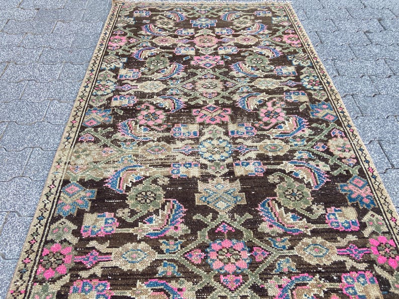 May include: A rectangular, vintage rug with a dark brown background and an intricate pattern. The design features floral and geometric elements in shades of pink, blue, and green. The rug has a distressed look, suggesting age and use.