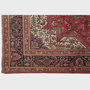 May include: A section of a traditional rug with a detailed, geometric pattern. The rug features a rich color palette of red, navy blue, cream, and touches of green and gold. The border is a complex design with repeating motifs.