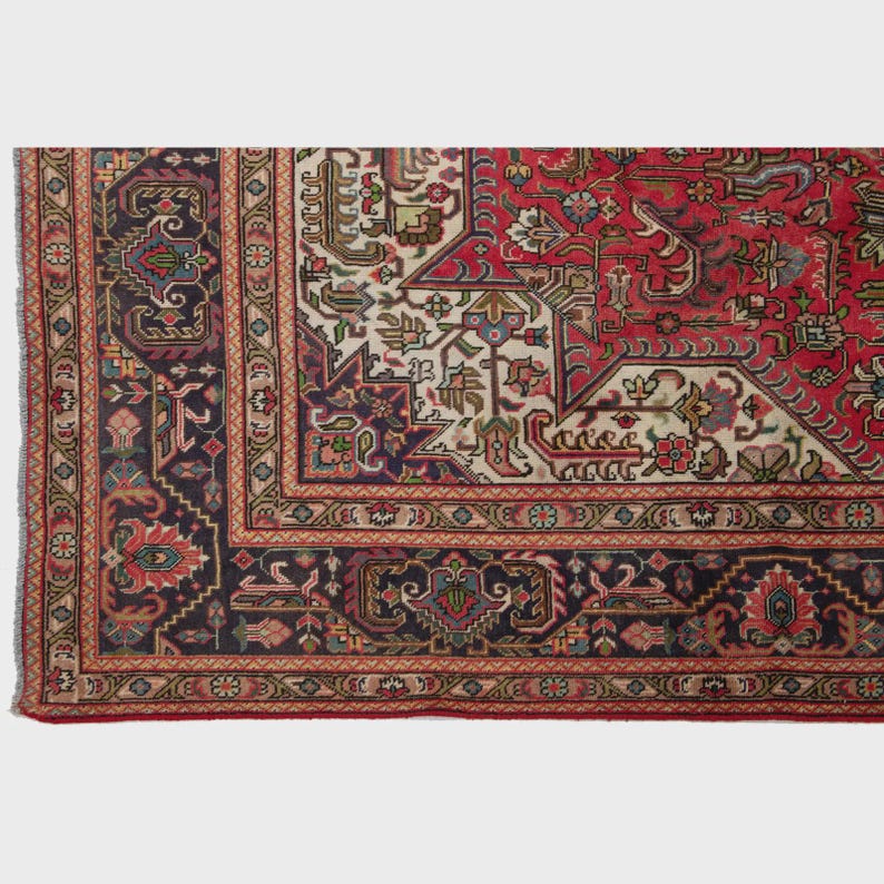 May include: A section of a traditional rug with a detailed, geometric pattern. The rug features a rich color palette of red, navy blue, cream, and touches of green and gold. The border is a complex design with repeating motifs.