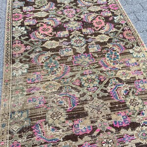 May include: A rectangular rug with an intricate pattern. The rug features a brown background with floral and geometric designs in shades of pink, blue, and beige. The rug's edges are bordered with a darker brown trim. The rug appears to be made of a woven material.