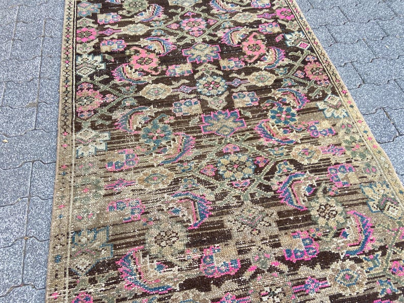 May include: A rectangular rug with an intricate pattern. The rug features a brown background with floral and geometric designs in shades of pink, blue, and beige. The rug's edges are bordered with a darker brown trim. The rug appears to be made of a woven material.