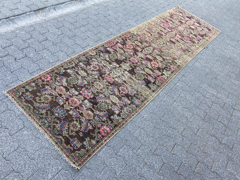 May include: A long, rectangular rug with a floral pattern in shades of brown, pink, blue, and green. The rug has a distressed, vintage look, with a faded appearance. The edges are fringed.