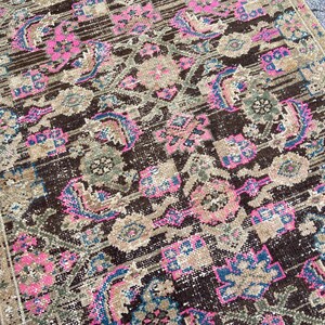 May include: A distressed, rectangular rug with a detailed, faded pattern. The rug features a dark brown central field with pink, blue, and green floral and geometric motifs. The border is a lighter tan color. The rug has a vintage, worn appearance.