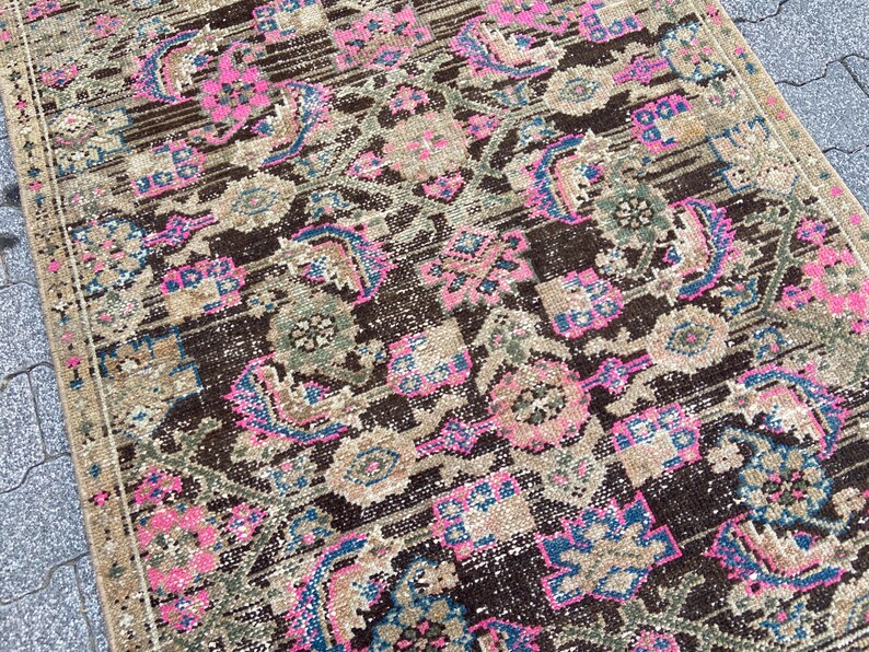 May include: A distressed, rectangular rug with a detailed, faded pattern. The rug features a dark brown central field with pink, blue, and green floral and geometric motifs. The border is a lighter tan color. The rug has a vintage, worn appearance.