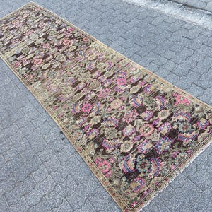 May include: A long, rectangular rug with an intricate floral pattern. The rug features a dark brown background with pink, purple, and green floral motifs. The edges are bordered with a beige trim. The rug is placed on a gray, textured surface.