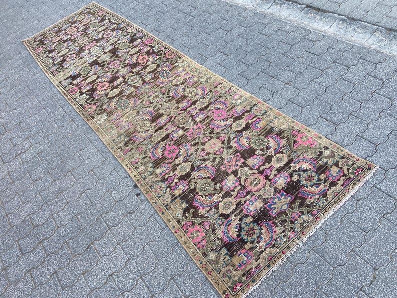 May include: A long, rectangular rug with an intricate floral pattern. The rug features a dark brown background with pink, purple, and green floral motifs. The edges are bordered with a beige trim. The rug is placed on a gray, textured surface.