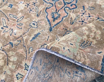 May include: A close-up of a vintage rug with a detailed floral pattern. The rug features a warm color palette of brown, blue, and peach. The edges of the rug are visible, showing the texture and construction of the rug.